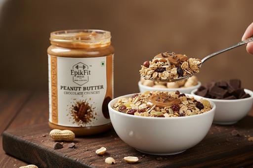 Jar of EpikFit Peanut Butter Chocolate Crunchy on a wooden surface with a bowl of granola and a spoon.